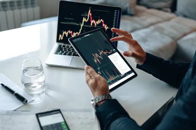 Close-up of a person using a tablet for online trading analysis with a laptop and glass of water nearby. Running social media agencies competitor research without paid tools